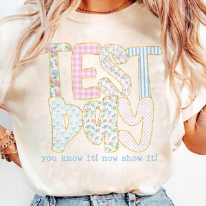May include: Cream-colored t-shirt with the words "TEST DAY" in a colorful, patchwork design. The letters are decorated with floral, gingham, and striped patterns. Below the text is the phrase "you know it! now show it!"