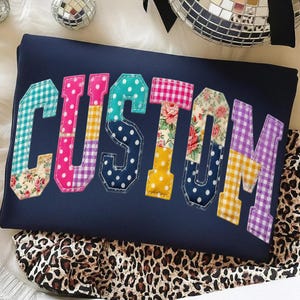 May include: Navy blue t-shirt with the word "CUSTOM" appliqued in colorful patchwork fabric. The letters feature various patterns, including polka dots, gingham, and floral prints. The shirt is resting on a leopard print fabric.