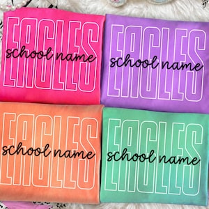 May include: Four folded t-shirts in bright pink, purple, orange, and green. Each shirt features the word EAGLES in large white outlined block letters and the text school name in black cursive script centered across the front of the garment.