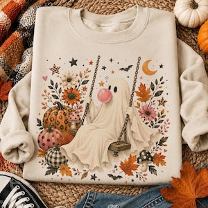 May include: A beige crewneck sweatshirt featuring a graphic print of a ghost sitting on a swing, blowing a pink bubble. The design includes colorful pumpkins, autumn leaves, flowers, mushrooms, stars, a crescent moon, and small bats.