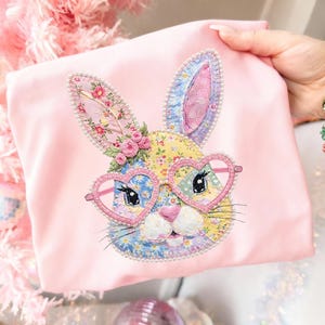 May include: A pink fabric item featuring an embroidered bunny face with heart-shaped glasses. The bunny has floral patterned ears and a patchwork face. The glasses are pink and the bunny is surrounded by a pearl-like trim.