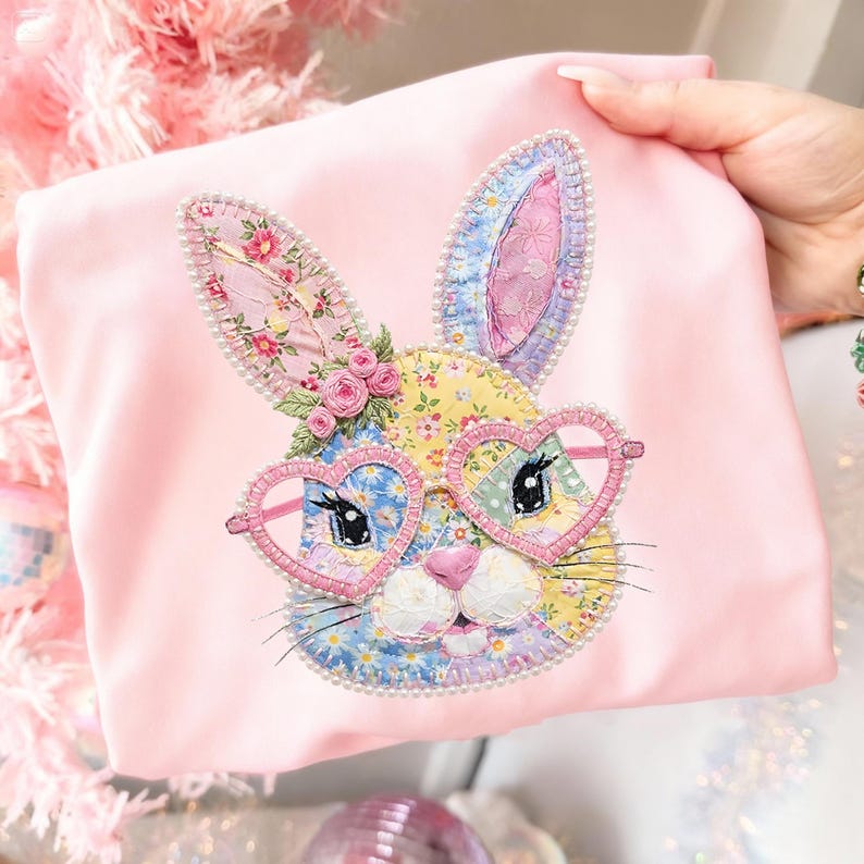 May include: A pink fabric item featuring an embroidered bunny face with heart-shaped glasses. The bunny has floral patterned ears and a patchwork face. The glasses are pink and the bunny is surrounded by a pearl-like trim.