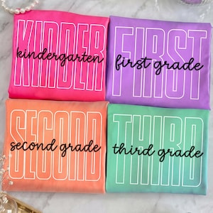 Könnte beinhalten: Vier gefaltete T-Shirts in leuchtenden Farben: Pink, Lila, Orange und Mintgrün. Jedes Shirt zeigt große, weiß umrandete Blockbuchstaben mit der Aufschrift KINDER, FIRST, SECOND und THIRD sowie die Wörter kindergarten, first grade, second grade und third grade in schwarzer Schreibschrift.