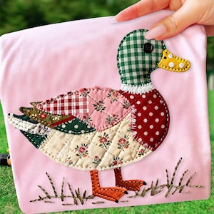 May include: A pink fabric square featuring a patchwork duck design. The duck has a green checkered head, a red polka-dot body, and wings with floral and patterned fabrics. Orange feet and embroidered grass complete the design.