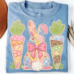 Puede incluir: Camiseta azul claro con un diseño de conejo de retazos y dos zanahorias. El conejo tiene un lazo rosa y estampados florales. Las zanahorias son verdes y marrones. El diseño está delineado con cuentas blancas. Una camiseta casual y cómoda.