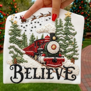 May include: A white square Christmas decoration featuring a detailed embroidered design. The design includes a red and green train, snow-covered trees, and the word "BELIEVE" in black script. Santa and his reindeer are flying in the sky.