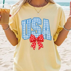 May include: A light yellow t-shirt with the word "USA" in blue gingham print. A red bow with white polka dots is centered below the text. The shirt is a casual style, suitable for patriotic events.