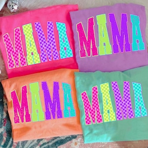Könnte beinhalten: Vier farbenfrohe T-Shirts, die jeweils das Wort "MAMA" in großen, fetten, karierten Buchstaben zeigen. Die Shirts sind in den Farbtönen Pink, Lila, Orange und Türkis gehalten. Die Buchstaben sind in verschiedenen Farben umrandet, was ein lebendiges und auffälliges Design ergibt.