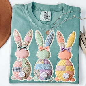 Könnte beinhalten: Hellblaugrünes Comfort Colors T-Shirt mit drei aufgenähten Hasen. Jeder Hase hat Patchworkmuster in Pastellfarben, eine Fliege und eine weiße Blume. Das Shirt hat einen Rundhalsausschnitt und ein kleines Etikett.