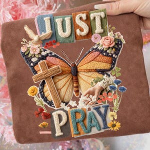Può includere: Quadrato marrone con ricamo di farfalla, croce e dettagli floreali. Il testo recita "JUST PRAY" in lettere strutturate. La farfalla ha ali arancioni, gialle e blu. È presente anche una colomba.