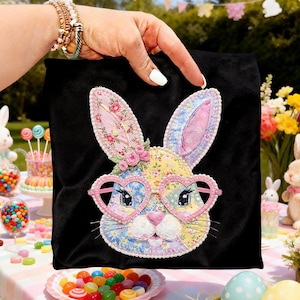 May include: A black square bag with a colorful appliqued bunny face. The bunny has heart-shaped glasses, floral ears, and a pink nose. The bag is held up by a hand, with a festive Easter-themed background.