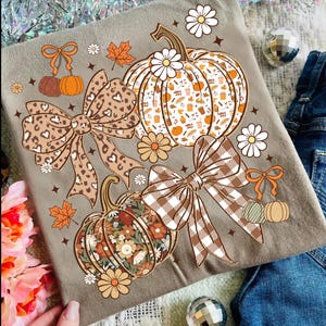 May include: A gray t-shirt featuring a fall-themed design. The design includes pumpkins, bows, fall leaves, and flowers in shades of brown, orange, and white. The shirt has a crew neck and short sleeves.