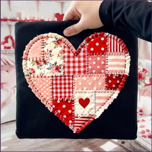 Pode incluir: Uma almofada quadrada preta com um grande design de coração de patchwork. O coração é composto por vários tecidos vermelhos e rosa, incluindo estampas florais, de bolinhas e xadrez. Um pequeno coração bordado está centrado no design.