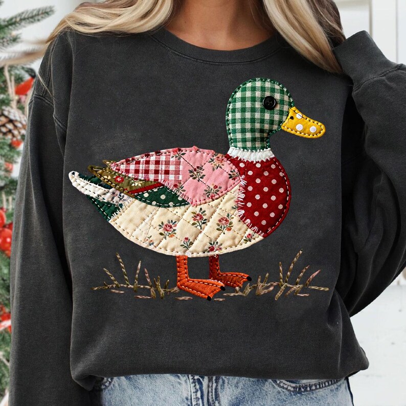 May include: Dark gray sweatshirt featuring a patchwork duck design. The duck has a green checkered head, a yellow dotted beak, and a body composed of various patterned fabrics in red, pink, and cream. The duck stands on embroidered grass.