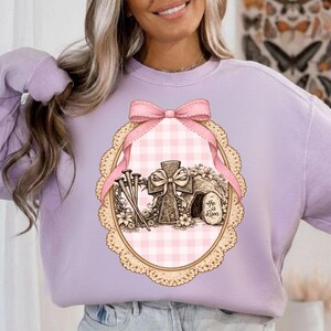 May include: A lilac sweatshirt with an Easter graphic. The design features a cross, nails, and a tomb with the text "He is Risen" within a pink and white gingham oval frame, decorated with a pink bow and lace edging.