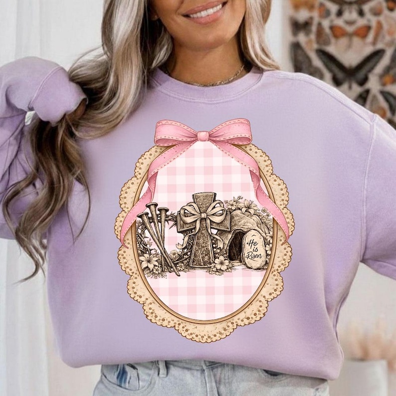 May include: A lilac sweatshirt with an Easter graphic. The design features a cross, nails, and a tomb with the text "He is Risen" within a pink and white gingham oval frame, decorated with a pink bow and lace edging.