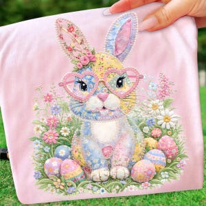 May include: A pink Easter-themed pillow featuring a patchwork bunny wearing heart-shaped glasses. The bunny is surrounded by colorful flowers and decorated Easter eggs. The pillow is held by a hand.