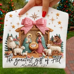 May include: Embroidered Christmas ornament featuring a Nativity scene with Mary, Joseph, and baby Jesus. The ornament has a pink bow, stars, and the text "The greatest gift of All" with "Matthew 1:21".