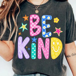 Op de afbeelding: Een antracietgrijs t-shirt met de tekst BE KIND in grote, kleurrijke, gedessineerde letters. De B is roze geblokt met hartjes, de E is blauw met madeliefjes, de K is paars met zebrastrepen, de I is groenblauw met witte hartjes, de N is roze met witte stippen en de D is geel met een smiley.