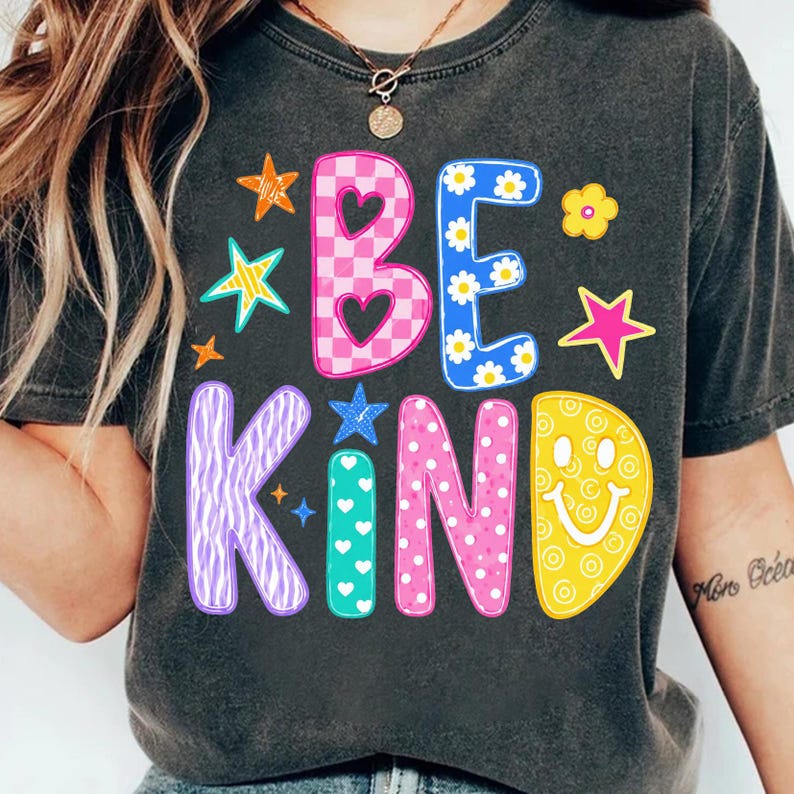 Op de afbeelding: Een antracietgrijs t-shirt met de tekst BE KIND in grote, kleurrijke, gedessineerde letters. De B is roze geblokt met hartjes, de E is blauw met madeliefjes, de K is paars met zebrastrepen, de I is groenblauw met witte hartjes, de N is roze met witte stippen en de D is geel met een smiley.