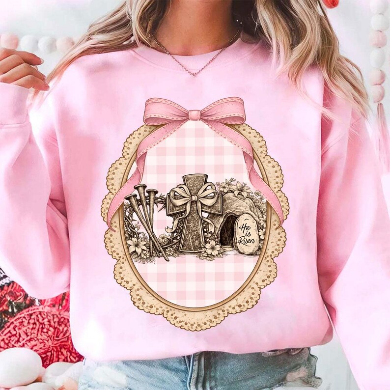 May include: Pale pink sweatshirt with an Easter graphic. The design features a cross, nails, and a tomb with the text "He is Risen" within an oval frame, a pink bow, and a gingham pattern.