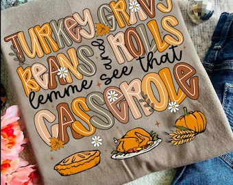 May include: A tan t-shirt with the text "Turkey Gravy Beans and Rolls lemme see that Casserole" in orange, brown, and white. The shirt features illustrations of a pie, turkey, pumpkin, and wheat.