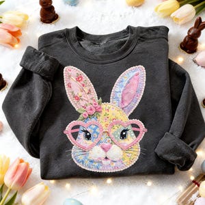 May include: Dark gray sweatshirt featuring a colorful bunny face applique. The bunny has heart-shaped glasses and floral accents. The design incorporates various patterns and colors, including pink, blue, and yellow. The sleeves are cuffed.
