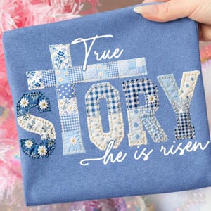 Può includere: Maglietta azzurra con le parole "True Story" e "he is risen" in scrittura bianca. Le lettere e la croce sono in stile patchwork con motivi floreali e a quadri blu. Una mano tiene la maglietta.