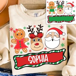 May include: Cream-colored sweatshirt featuring Christmas-themed graphics. The design includes a gingerbread woman, a reindeer with a red bow, and Santa Claus. The name "SOPHIA" is displayed in white on a red banner with a green border. Additional graphics include snowflakes.