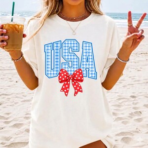 May include: White t-shirt with the word "USA" in blue gingham print and a red bow with white polka dots. The shirt is a casual style, perfect for patriotic events.