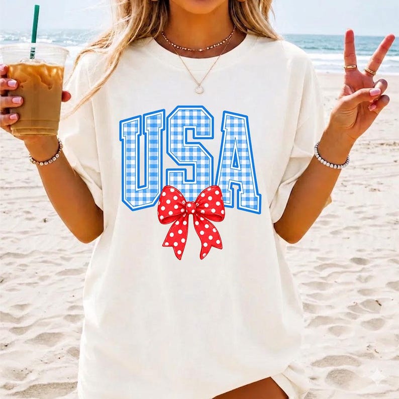 May include: White t-shirt with the word "USA" in blue gingham print and a red bow with white polka dots. The shirt is a casual style, perfect for patriotic events.