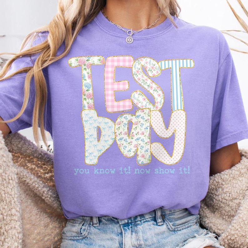 May include: A light purple t-shirt with the words "TEST DAY" in a patchwork design. The letters are decorated with floral, gingham, and striped patterns. Below the text is the phrase "you know it! now show it!"