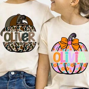 May include: Two cream-coloured t-shirts with pumpkin designs. One shirt features a pumpkin with a baseball cap and the name "Oliver" in brown and green. The other has a pumpkin with a bow and the name "Olivia".