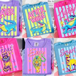 Può includere: Collezione di magliette a maniche lunghe colorate con grafiche di animali e testo. I disegni includono un alligatore con occhiali da sole, un procione, una tigre e un asino. Ogni maglietta presenta una frase unica come "LATER" o "EASY" in grassetto.