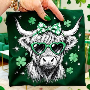 May include: A dark green t-shirt featuring a black and white illustration of a Highland cow wearing heart-shaped sunglasses and a bow with shamrocks. The shirt has shamrock and star accents.