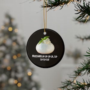 May include: A round black Christmas ornament with a white mozzarella ball topped with a green basil leaf. The ornament has the text "MOZZARELLA-LA-LA-LA, LA-LA-LA-LA!" printed on it.