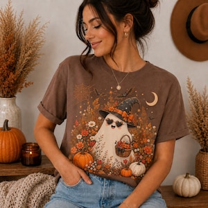Peut inclure: Un t-shirt marron à manches courtes orné d'un motif représentant un fantôme blanc portant un chapeau de sorcière noir avec un nœud orange et des lunettes en forme de cœur. Il tient un panier de citrouilles parmi des fleurs d'automne et des chauves-souris.