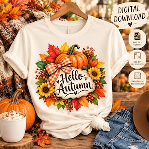 Może przedstawiać: Kremowy t-shirt z grafiką przedstawiającą dynię, słoneczniki, czerwone jagody i jesienne liście ułożone w wieniec. Na dyni znajduje się pomarańczowo-biała kokarda w kratkę. Napis Hello Autumn jest wykonany czarną czcionką.