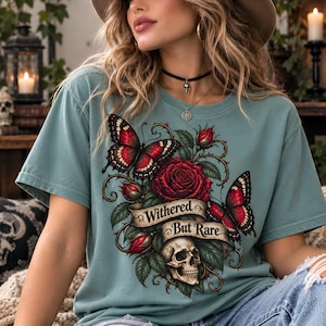 Peut inclure: Un t-shirt vert sauge avec une impression graphique représentant deux papillons rouges et noirs, une grande rose rouge avec des boutons et un crâne humain. Une bannière en ruban beige affiche le texte noir Withered But Rare en police serif.