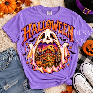 May include: A lavender short-sleeve t-shirt featuring a large graphic of a ghost with a barn, pumpkins, and bats inside its body. The word HALLOWEEN is printed in large orange letters above the ghost. The shirt has a crew neckline and short sleeves.