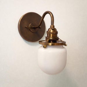 May include: A vintage-style wall sconce with a round, bronze-colored base and curved arm. The fixture features a white, globe-shaped glass shade and brass-colored accents. The design evokes an industrial or farmhouse aesthetic.