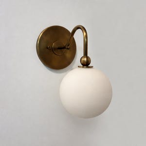 Puede incluir: Una lámpara de pared con una placa trasera redonda de color latón y un brazo curvo. La lámpara presenta una pantalla esférica de vidrio blanco. El diseño es simple y elegante, adecuado para varios estilos de interiores.