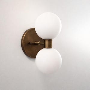 May include: A wall sconce featuring a circular antique brass backplate and a horizontal arm that supports two stacked, spherical white frosted glass globes. The metal components have a warm, metallic finish, while the glass shades are opaque.