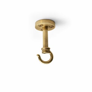 May include: A brass ceiling hook with a circular base and a curved hook. The hook is attached to a cylindrical post, and the base is secured with three screws. The hook is designed for hanging items from the ceiling.