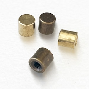 May include: Four small, cylindrical metal objects in varying shades of gold and bronze are scattered on a white surface. One has a threaded interior, while the others are solid or hollow. The objects appear to be hardware components.