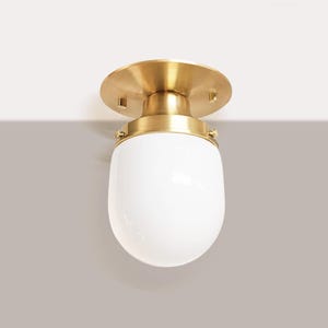 May include: A flush mount ceiling light fixture with a brass finish and a white, teardrop-shaped glass shade. The fixture is mounted on a white ceiling and has a simple, elegant design. The light is suitable for various interior styles.