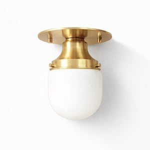 May include: A brass ceiling light fixture with a round base and a white, rounded glass shade. The fixture has a classic design, with a polished brass finish and a clean, modern aesthetic. The light is mounted on a white wall.