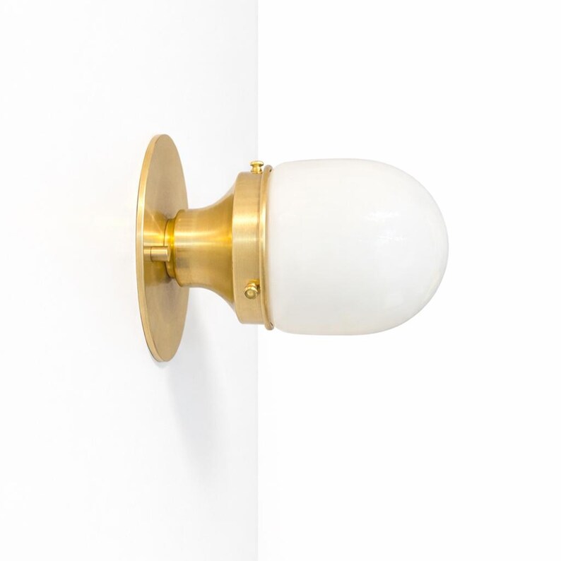 May include: A wall-mounted light fixture with a round brass base and a cylindrical brass neck. The fixture features a white, oval-shaped glass shade. The light is designed to be mounted on a wall.