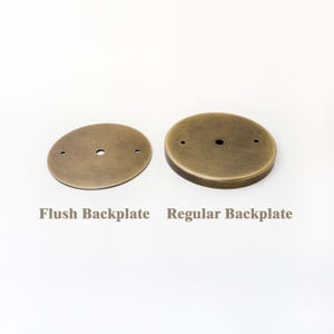 May include: Two circular metal backplates with an antique brass finish. The left item is labeled Flush Backplate and is flat, while the right item is labeled Regular Backplate and features a raised, rounded edge. Both have central holes.