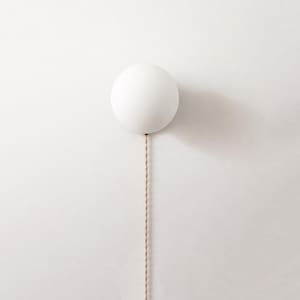 May include: A white, spherical wall sconce with a beige cord. The sconce is mounted on a white wall, casting a shadow to the right. The design is minimalist and modern, suitable for interior lighting in a home.